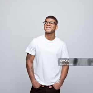 Happy african young man wearing white t-shirt and eyeglasses, standing with hands in pockets, looking away and smiling. Studio portrait on grey background.