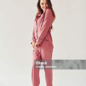 Cheerful young woman in pajamas standing indoors against white background in the studio.