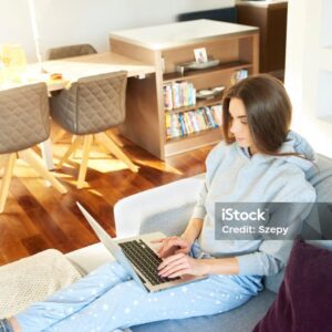 High angle shot of young woman wearing hoodie and relaxing on sofa while using her laptop at home.